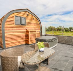 An outdoor patio with a wooden cabin hot tub wicker chairs and a glass table with flowers at Caban Cariad in Bryngwran near Bodedern