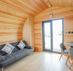 A wooden room with a gray sofa with pillows a round table with chairs and a door at Caban Cariad in Bryngwran near Bodedern