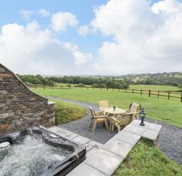 An outdoor patio with a hot tub and wooden table and chairs on gravel with a fenced grassy field in the background at Tan Y Wawr - Derwen in Llanfair Caereinion