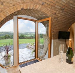 A room with a round brick ceiling and a glass door open to a stone patio with green fields beyond and a countertop with a bottle and four wine glasses at Tan Y Wawr - Derwen in Llanfair Caereinion