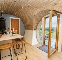 A kitchen with a curved brick ceiling wooden stools a countertop with a fruit basket and champagne bottle and an open door leading outside at Tan Y Wawr - Derwen in Llanfair Caereinion