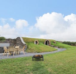 An outdoor area with a wooden table and chairs next to a stone wall and hot tub and a house built into a grassy hill at Tan Y Wawr - Derwen in Llanfair Caereinion