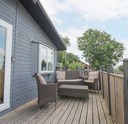 An outdoor wooden deck with wicker chairs sofa and table attached to a house at Wayside Lodge in Aston On Clun