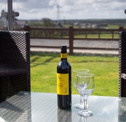 A glass table with a bottle of wine and two empty wine glasses between two wicker chairs on a patio overlooking a lawn at The Lodge in Rhosybol