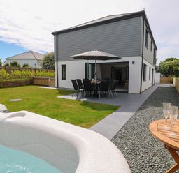 A backyard with a hot tub a patio table and chairs under an umbrella and a wooden table with glasses and a champagne bucket at Tredarn in Connor Downs
