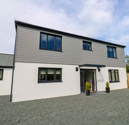 A modern two-story house with grey cladding on the upper floor white walls on the lower floor black framed windows and a gravel front yard at Tredarn in Connor Downs