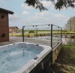 An outdoor patio with a hot tub next to a wooden building and a pond with trees at Ruperts Retreat in Nateby near Garstang