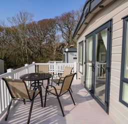 An outdoor deck with a glass table and four chairs attached to a beige mobile home with white railing and trees in the background at Crosswinds 68 in Bembridge