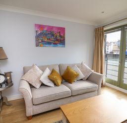 A living room with a beige sofa with cushions a wooden coffee table a side table with a lamp clock and flowers and large glass doors at Flat 2 in Pembroke