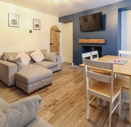 A living room with beige sofas a wooden dining table with white chairs a wall-mounted tv and a fireplace at Gwel Y Mor in Beaumaris
