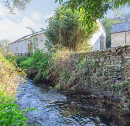 A stream flowing beside a stone wall with plants and two buildings in the background at Millers Lodge in St Keyne near Liskeard