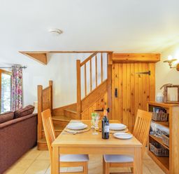 A dining area with a wooden table set for two next to a wooden staircase and a bookshelf at Millers Lodge in St Keyne near Liskeard