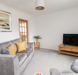 A living room with a gray sofa with cushions a wooden tv stand with a television a side table with a plant and a glass door at Sandpiper in Weymouth