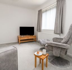 A living room with a grey armchair and footstool a wooden side table with a plate and mug a wooden TV stand with a TV and a grey sofa at Sandpiper in Weymouth
