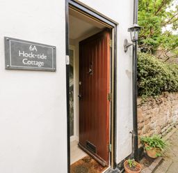 An open wooden front door with a doormat next to a wall sign reading Hock-tide Cottage and a stone wall with plants at Hock-tide Cottage in Kenilworth
