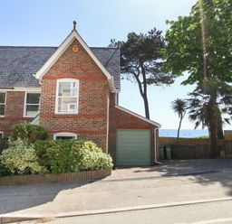 A brick house with a green garage door and bushes in front near trees and a view of the sea at Castle Cove Cottage in Weymouth