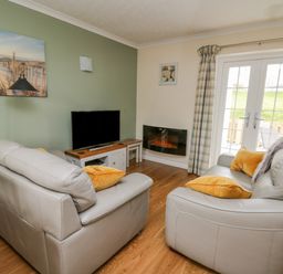 A living room with two leather sofas yellow cushions a television an electric fireplace and double doors with curtains at Oak Tree Cottage in Aberporth