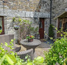 An outdoor patio with wicker chairs a table with flowers and a wooden bench against a stone wall at Nightingale Cottage in Lostwithiel