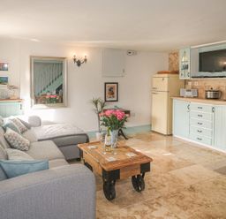 A living room with a grey sofa wooden coffee table and kitchen area with pastel cabinets at Nightingale Cottage in Lostwithiel