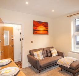 A small living room with a brown sofa and ottoman by a window and a wooden dining table with plates at Pams House in Weymouth