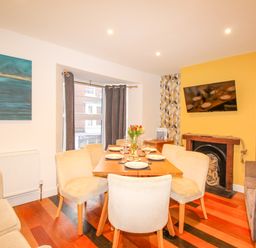 A dining room with a wooden table set for six people a sofa a wall-mounted TV and a window with curtains at Pams House in Weymouth