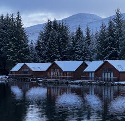 Three cabins with snow on roofs by a lake surrounded by pine trees and mountains at Ptarmigan Lodge in Glengoulandie near Aberfeldy