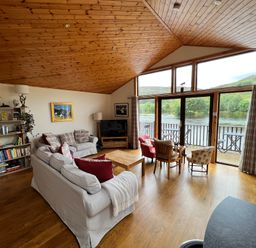 A living room with sofas a bookshelf large windows and a view of a river at Ptarmigan Lodge in Glengoulandie near Aberfeldy