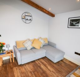 A living room with a grey sectional sofa with cushions a wooden side table with flowers and lamp and wall art at Celyn in Cyffylliog