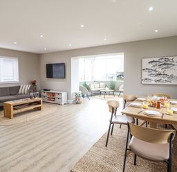An open living and dining area with a gray sofa, wooden coffee table, wall mounted TV, dining table set with plates and glasses, and a seating nook at Bryn Hafod in Dyffryn Ardudwy