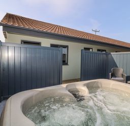 An outdoor patio area with a hot tub a chair a table with glasses and a bottle behind a grey fence at Chalet 1 in Sherburn