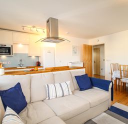 An open plan living area with a beige sofa with blue pillows a wooden dining table with chairs and a kitchen with white cabinets and appliances at Broadleys in Swanage