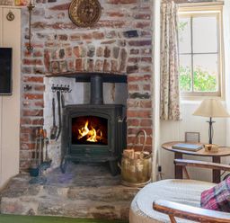A room with a lit wood stove set in a brick fireplace a wooden chair with a cushion a small table with a lamp and a window at End Cottage in Malborough