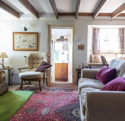 A living room with a beige sofa with purple cushions an armchair a red patterned rug and a window seat at End Cottage in Malborough