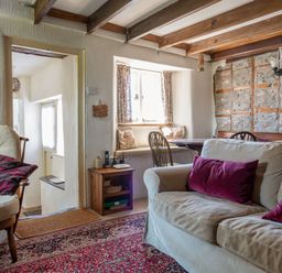 A living room with a beige sofa with red pillows a wooden chair a small wooden side table a window with curtains and a dining area at End Cottage in Malborough