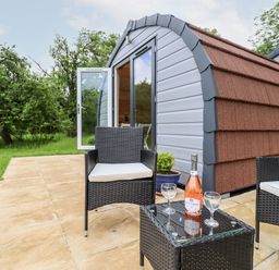 A patio with two wicker chairs and a small table with a bottle and glasses outside a small cabin at Meadow Lodge @ Berrys Place Farm in Churcham