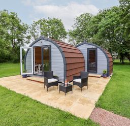 Two small curved lodges with doors and windows on a paved patio with two chairs and a table at Meadow Lodge @ Berrys Place Farm in Churcham