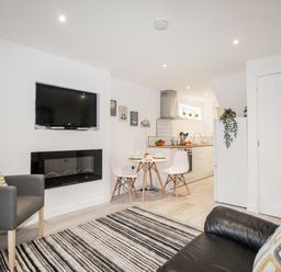 A living room with a striped rug a wall-mounted TV a small dining table with two white chairs and a kitchen area at Portside Greenhill Weymouth