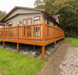 A wooden cabin with a raised deck and railing next to a paved driveway and grass at 16 Heritage Park in Heritage Park near Kilgetty