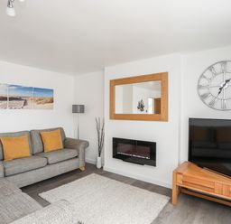 A living room with grey sofas yellow cushions a wooden TV stand and a wall clock at 31 Beach Road Morfa Bychan