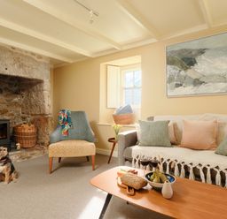 A living room with a stone fireplace a chair a sofa cushions a wooden coffee table with fruit and bread and a small dog on the floor at Faraway Cottage in St Levan near Porthgwarra