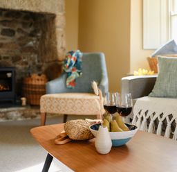 A living room with a coffee table holding bread pears and wine glasses by a stone fireplace at Faraway Cottage in St Levan near Porthgwarra