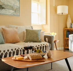 A living room with a sofa, pillows, a wooden coffee table with bread, cheese, two glasses of red wine, pears, and a small vase at Faraway Cottage in St Levan near Porthgwarra