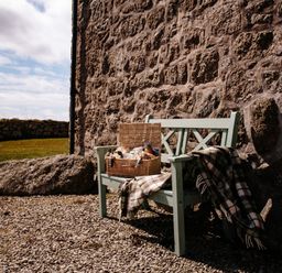 A green wooden bench with a picnic basket and a plaid blanket beside a stone wall outdoors