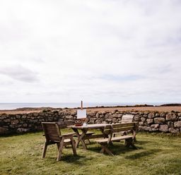 An outdoor wooden table with chairs and an easel on grass next to a stone wall with a field and ocean in the background at Three Chimneys in St Levan near Porthgwarra