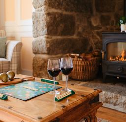 A living room with a Scrabble game and two glasses of wine on a wooden table near a lit fireplace at Three Chimneys in St Levan near Porthgwarra