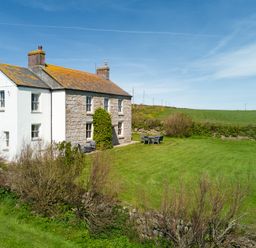 A stone and white painted house with two chimneys and garden furniture on a lawn at Higher Roskestal in St Levan near Porthgwarra