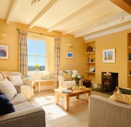 A living room with sofas a wooden coffee table flowers on the table a window with curtains and a fireplace at Higher Roskestal in St Levan near Porthgwarra
