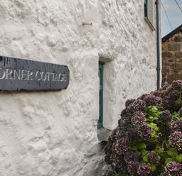 A white textured wall with a sign saying corner cottage next to a bush with purple flowers at Corner Cottage in Porthgwarra