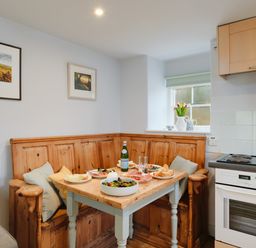 A kitchen dining area with a wooden corner bench and table set with food and drinks at Corner Cottage in Porthgwarra