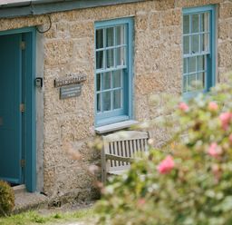 A stone cottage exterior with a blue door and windows a wooden bench and a flower bush at Cove Cottage in Porthgwarra